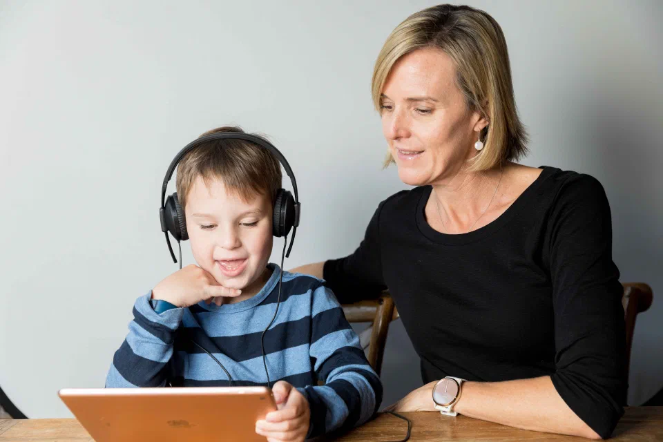 Child wearing headphones looking at an iPad while parent supervises.