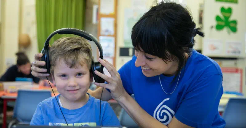 Young boy smiling at the camera while a lady sitting next to him wearing a Sound Scouts t-shirt places headphones over his ears.