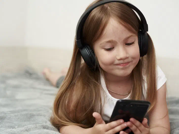 Young girl lying on her bed holding a mobile device while wearing wireless headphones.