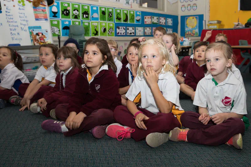 A group of young students sitting cross-legged on the floor of a classroom looking up and listening to something off-screen.