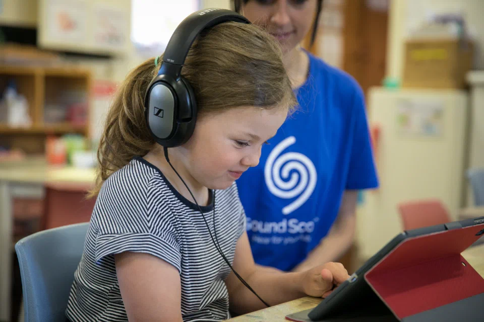 A young child wearing headphones looking at an iPad with an adult in the background wearing a Sound Scouts shirt.