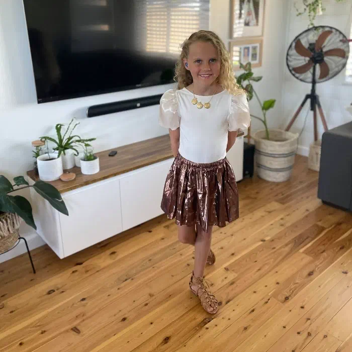 Laila standing in her living room smiling, feet-crossed, hands behind her back sporting her blue in-ear hearing aid.